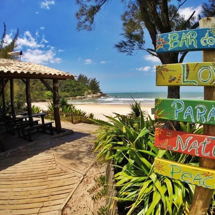 Uma placa de madeira com tábuas coloridas pintadas à mão indica a direção de uma praia de areia com um pátio coberto sob um céu claro.