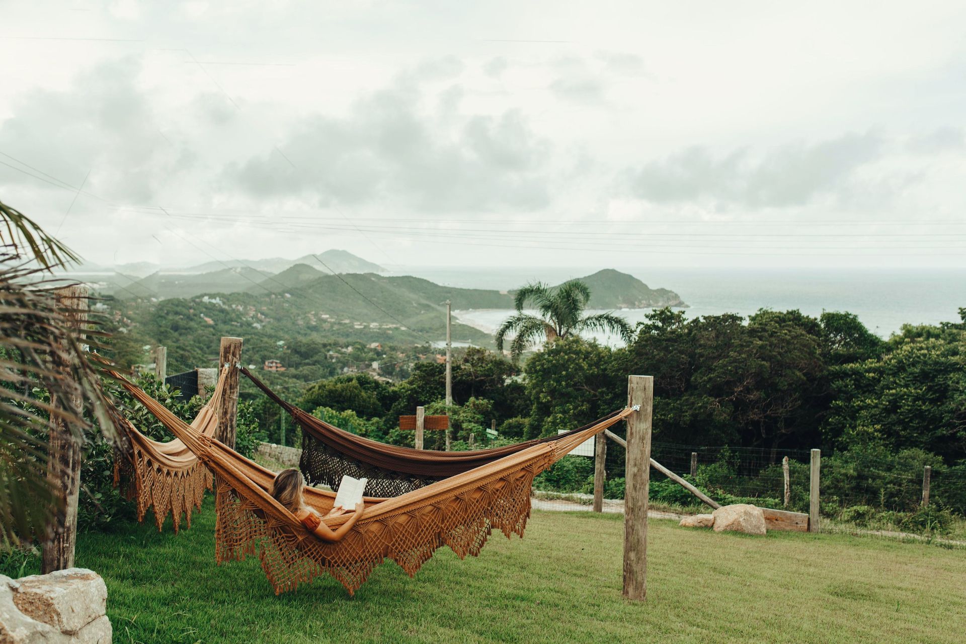 Uma pessoa relaxa em uma rede trançada ao ar livre, com vista para uma paisagem exuberante e montanhosa e para o oceano distante sob um céu nublado.