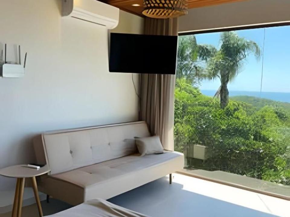 Um quarto moderno e luminoso com um sofá bege, uma pequena mesa lateral, uma TV fixada na parede e vista para as árvores e para o oceano.