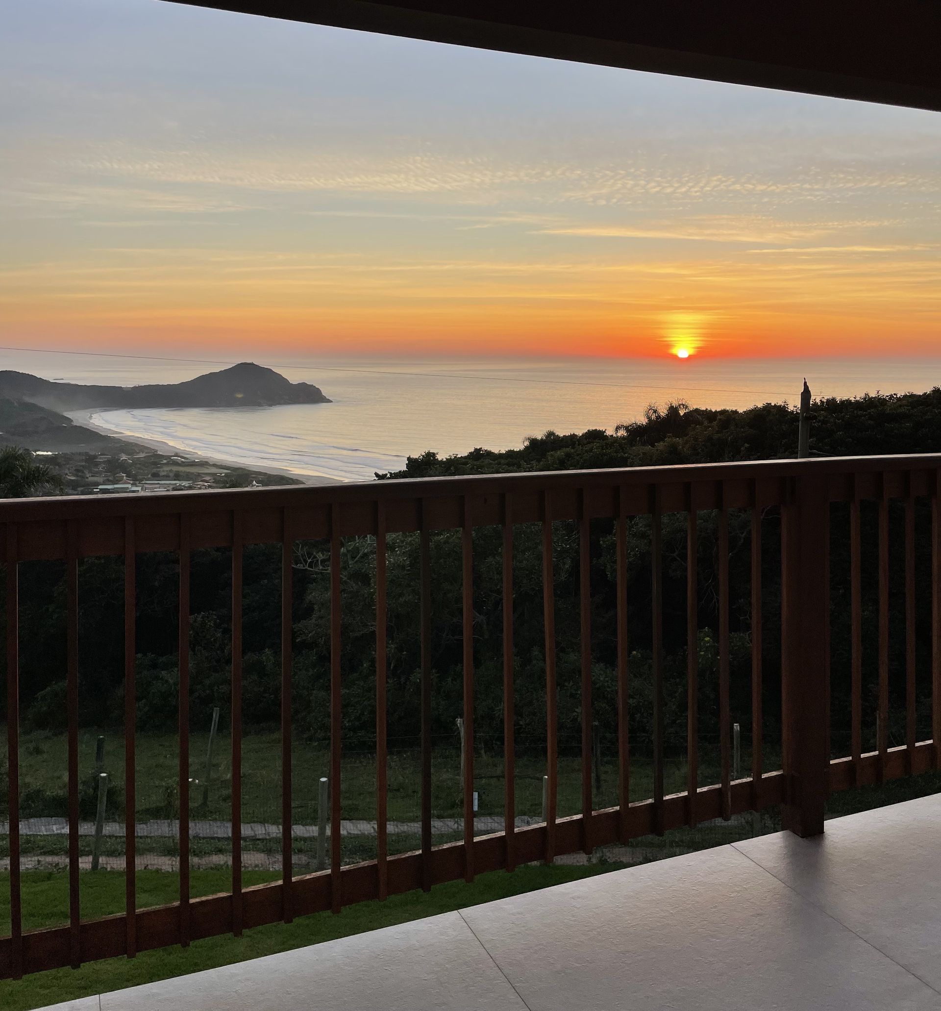 Uma balaustrada de madeira oferece vista para uma paisagem costeira ao pôr do sol, com o sol dourado se pondo sobre o oceano e um promontório distante.