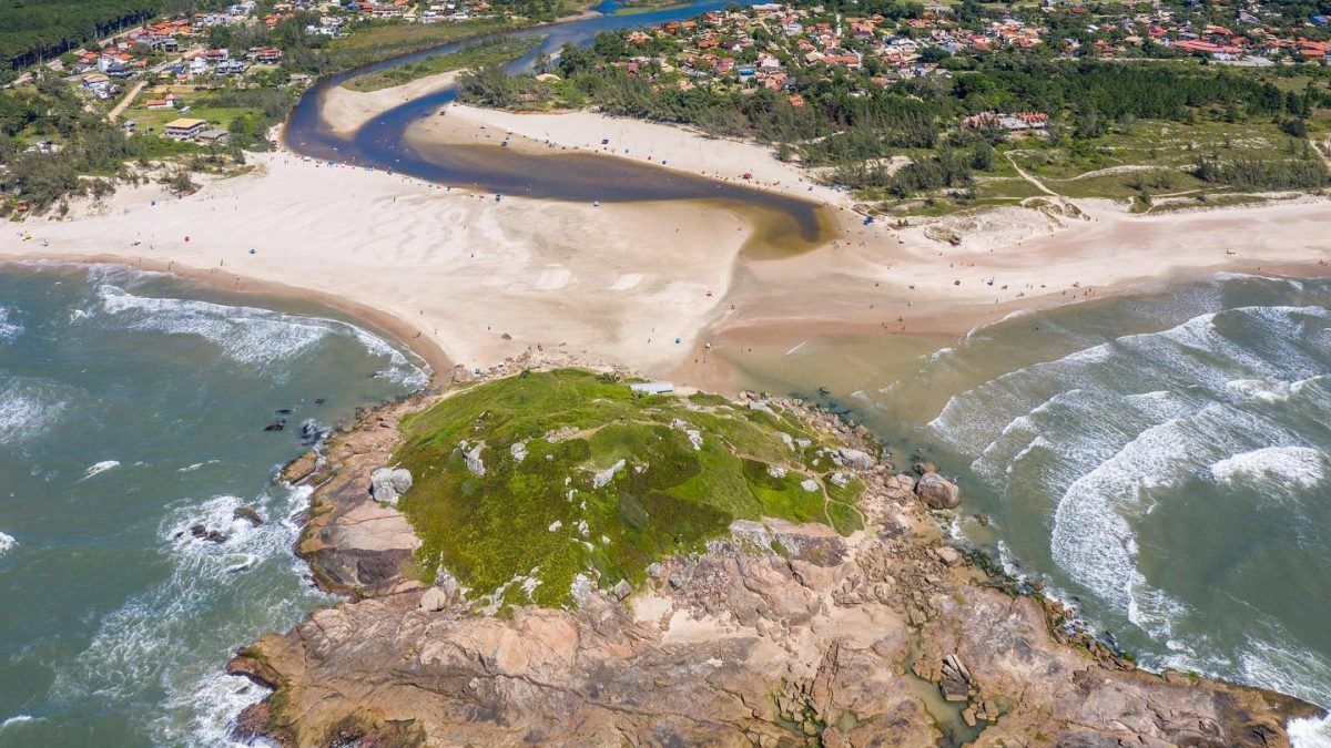 Vista aérea da foz de um rio encontrando o oceano, com um promontório rochoso verde dividindo a praia de areia da água costeira.