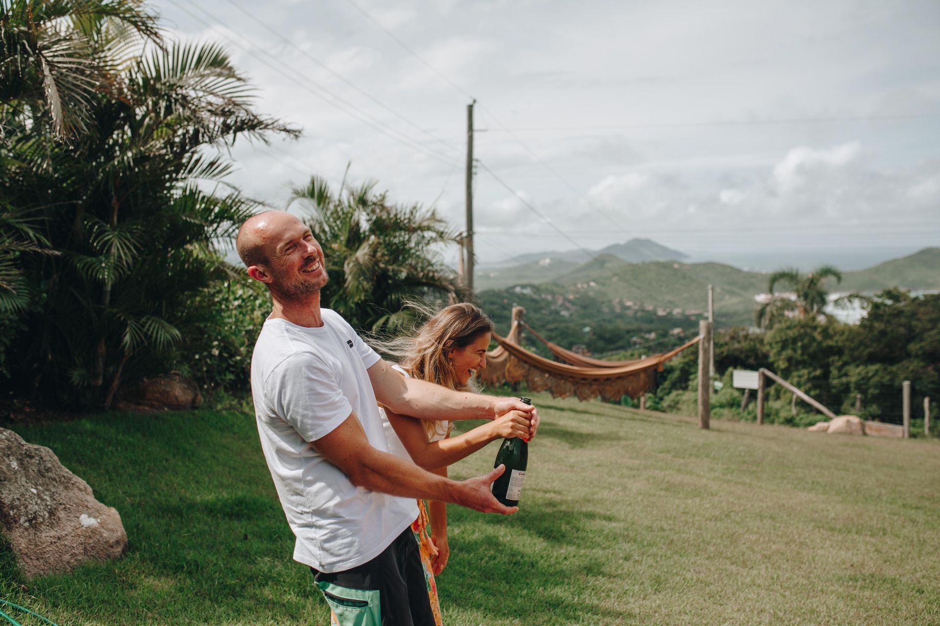 Uma pessoa sorridente estoura uma garrafa de champanhe ao ar livre, em uma encosta gramada com palmeiras e colinas distantes ao fundo.
