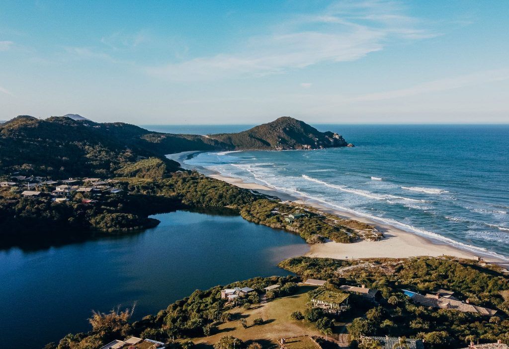 Vista aérea de um lago azul escuro que encontra uma praia de areia, cercado por colinas verdejantes e o oceano sob um céu azul.