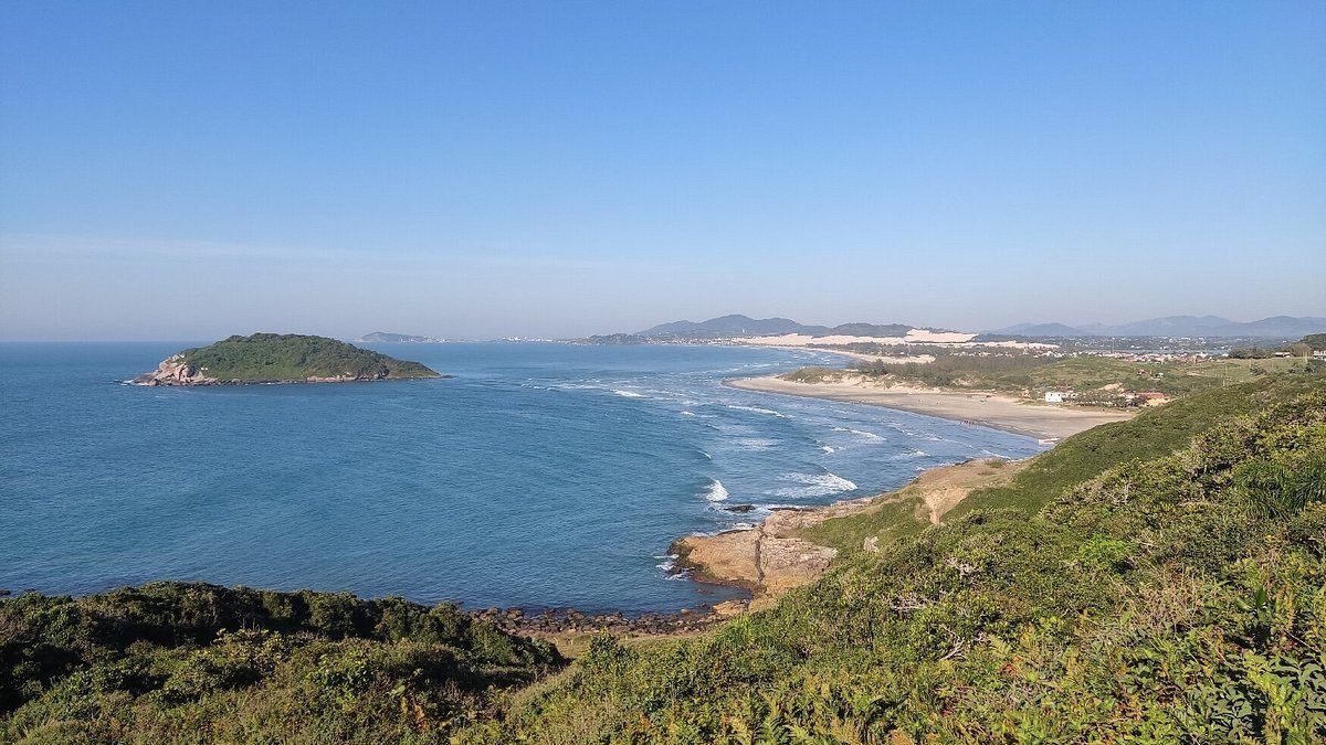 Uma vista costeira com uma pequena ilha em um mar azul calmo, praia de areia e uma costa verdejante e ondulada sob um céu claro.