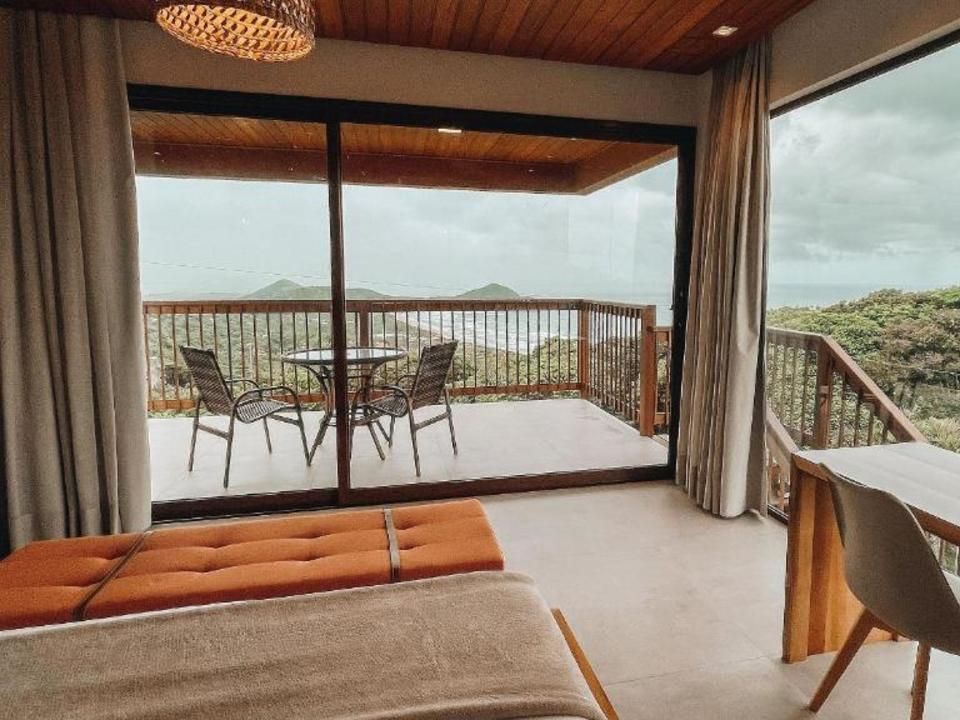 Vista de um quarto de hotel através de grandes portas de correr para um terraço com duas cadeiras, com vista para a floresta e o oceano.