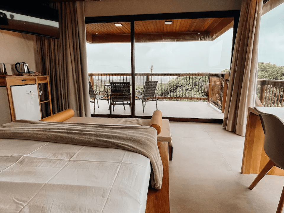 Um quarto de hotel com uma cama grande em primeiro plano, portas de vidro deslizantes que se abrem para uma varanda com assentos e vista para a natureza.