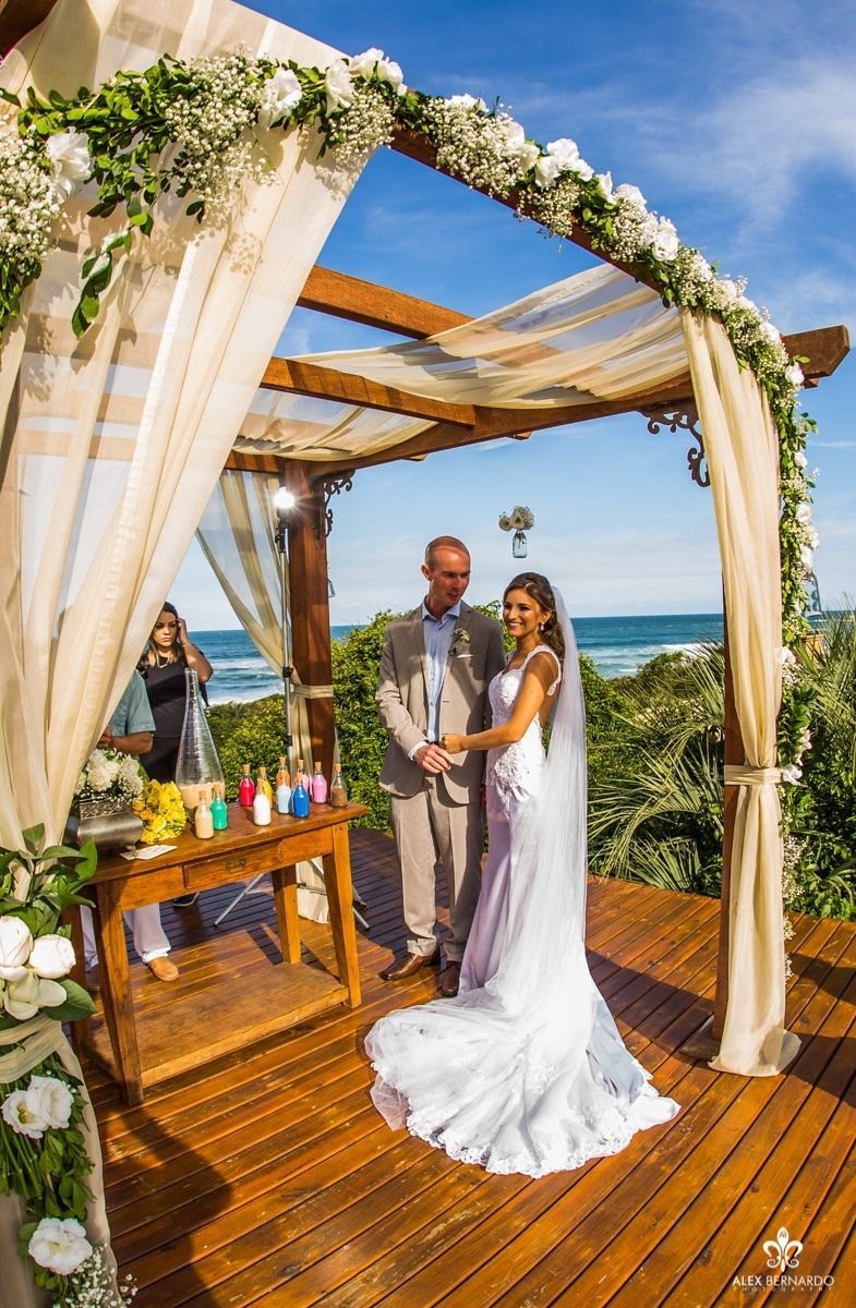 Um casal está de pé sob um arco de madeira decorado com flores, em um deck com vista para o oceano, em um dia ensolarado.
