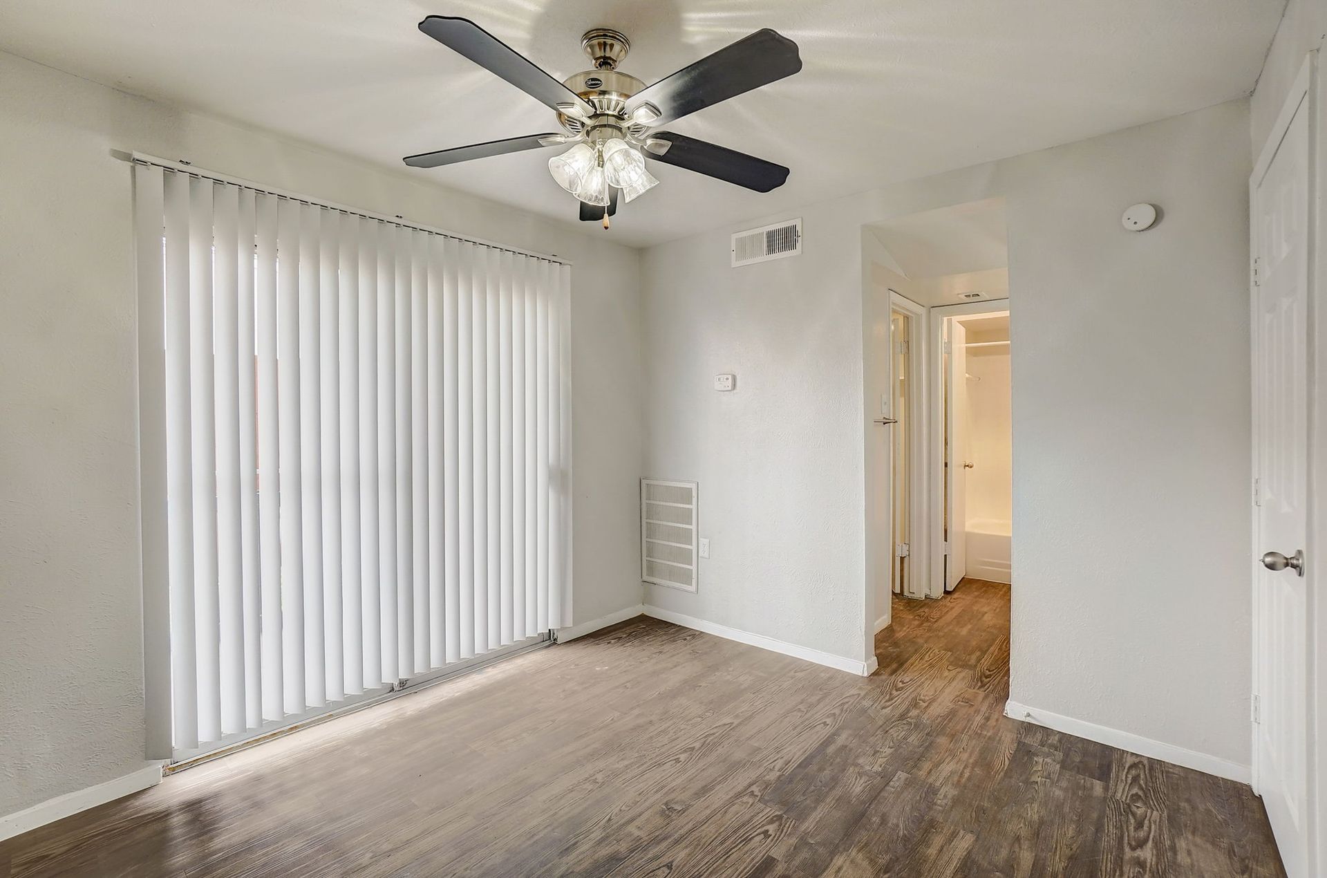 Empty room with a ceiling fan, sliding door with vertical blinds, and a doorway to a bathroom.
