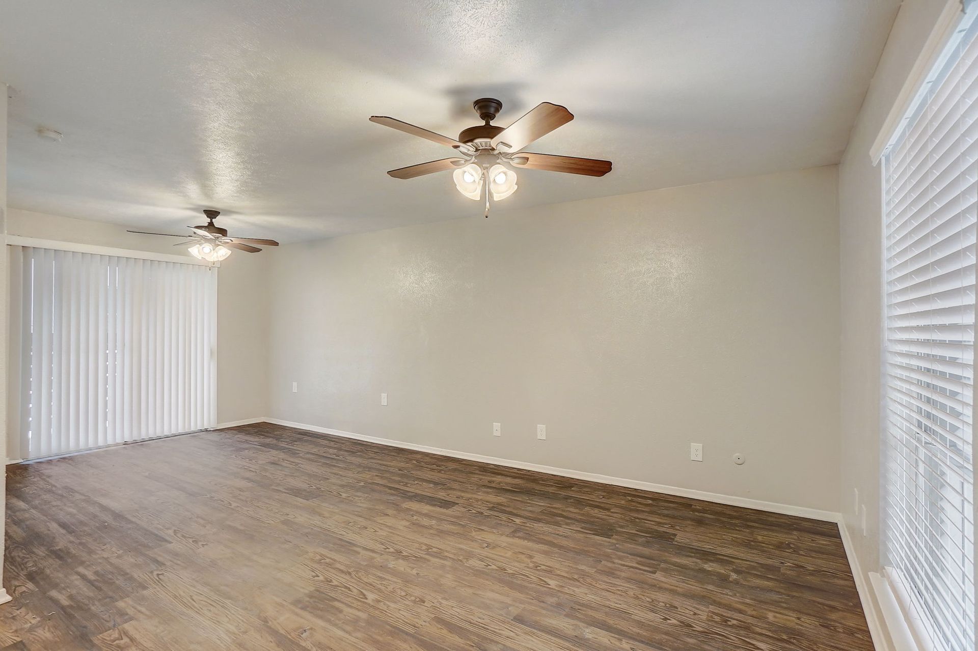 Empty living room with hardwood floors, white walls, two ceiling fans, and a sliding glass door.