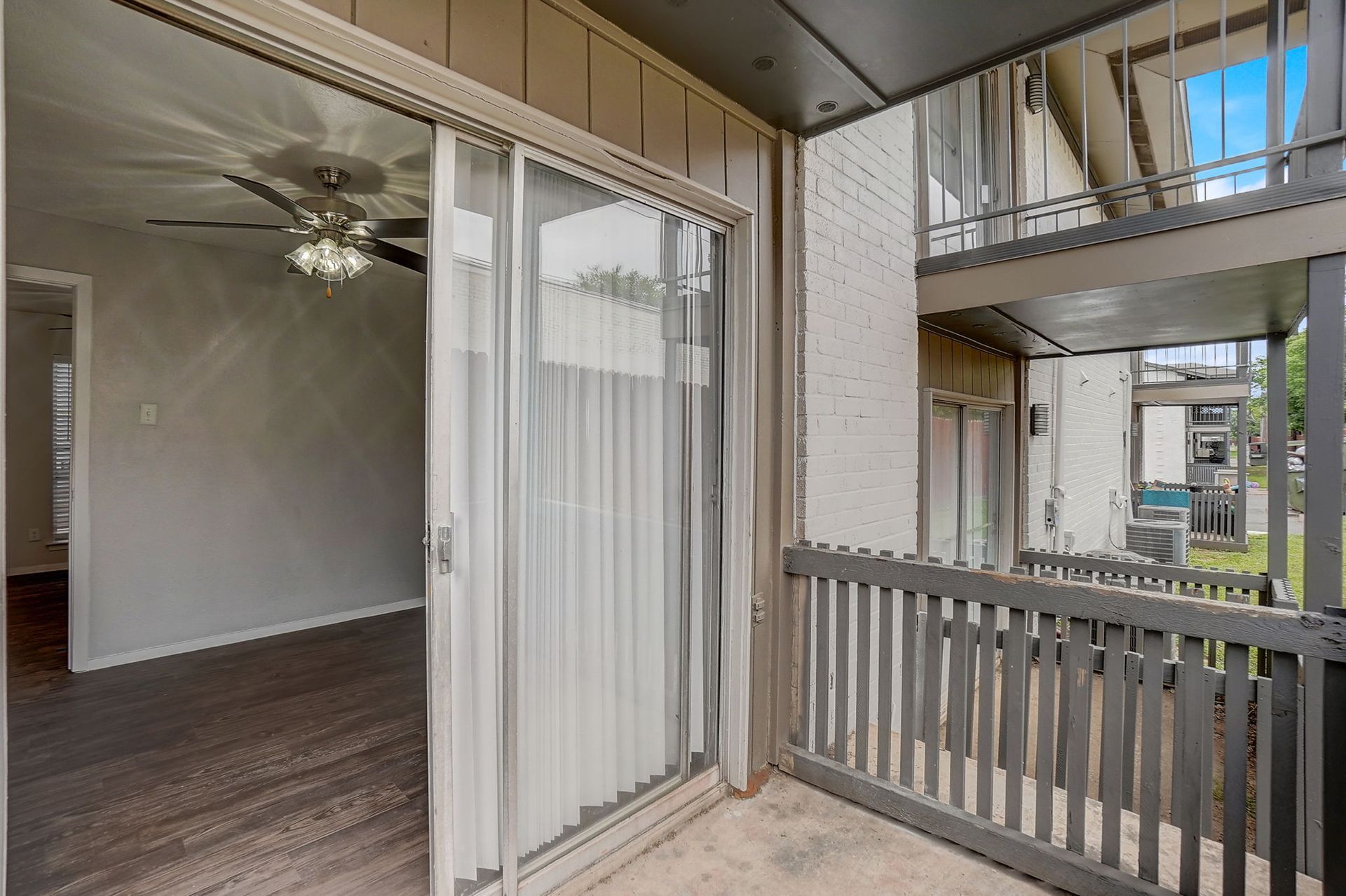Balcony with sliding glass door open to interior room. Gray railing, white curtains, and building exterior visible.
