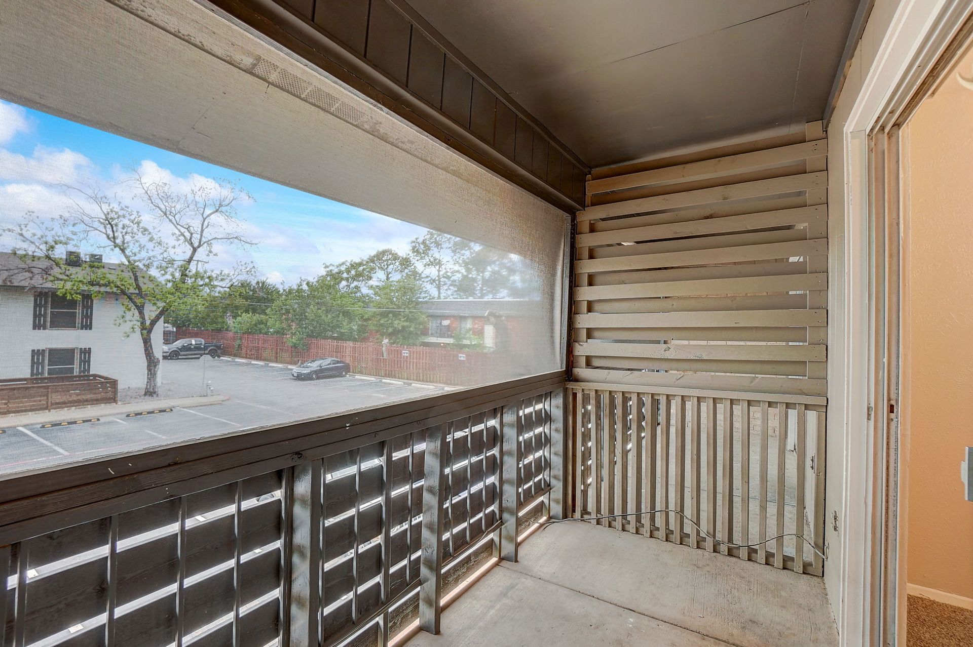 Balcony overlooking a parking lot and trees. Wooden railing and wall with an open door.