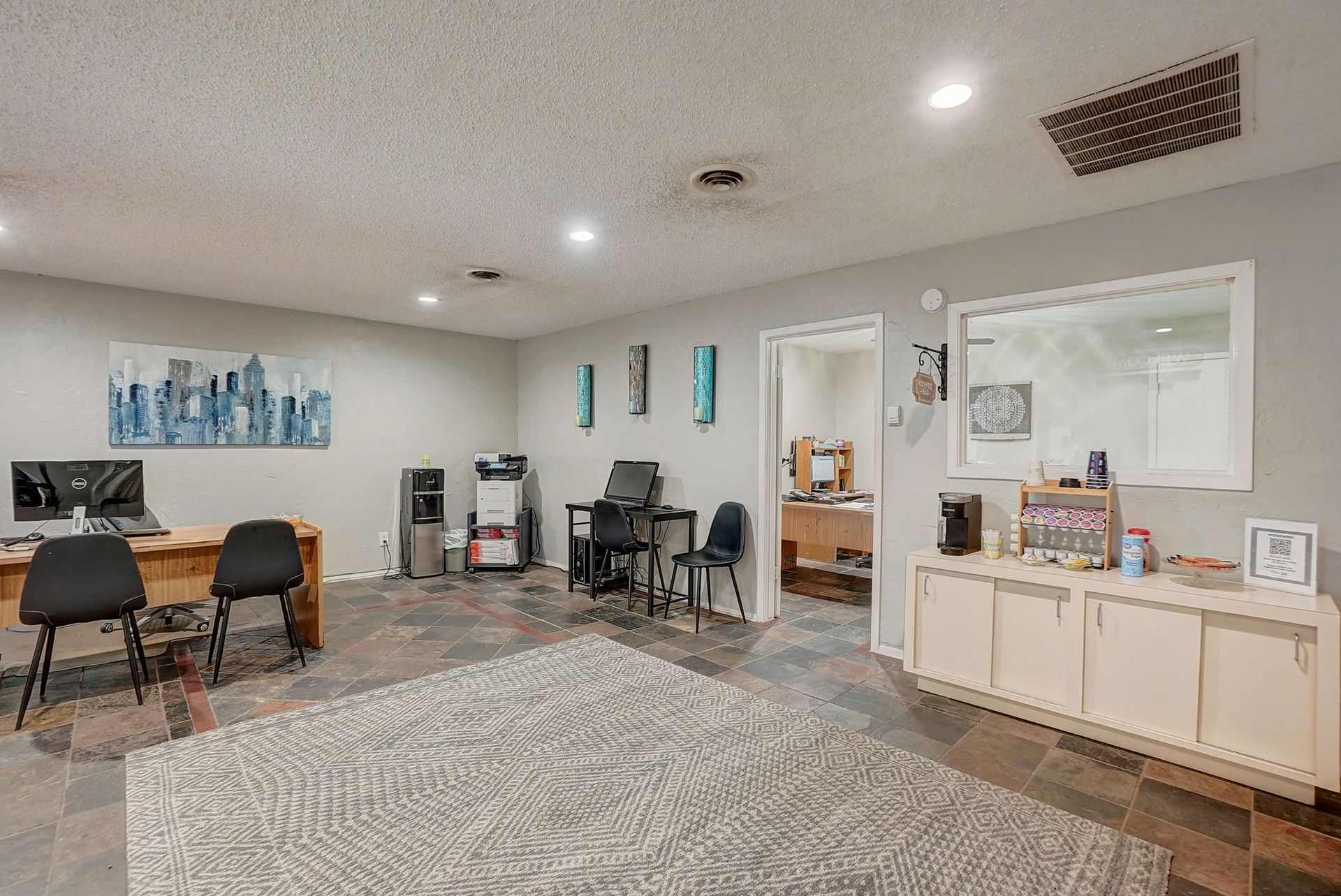 Office interior with desks, computers, chairs, rug, and cabinets. Light gray walls, recessed lighting.