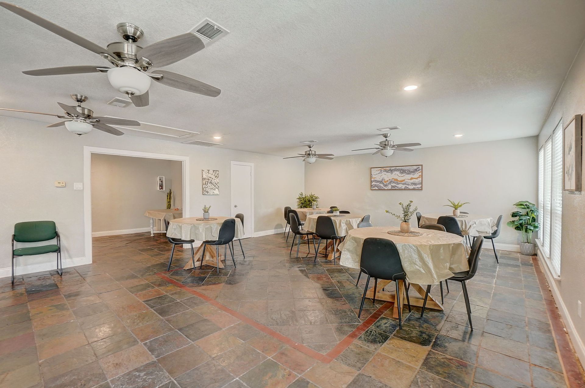 Dining room with round tables, draped in tablecloths, black chairs, slate floor, ceiling fans.