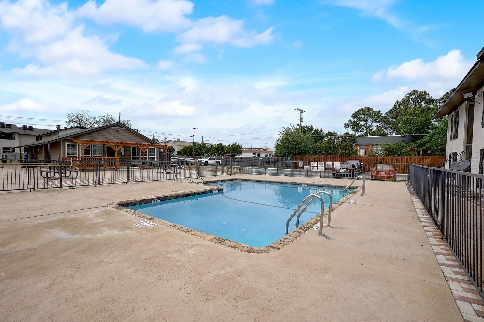 Swimming pool surrounded by concrete, near a building with a pergola, under a blue sky.