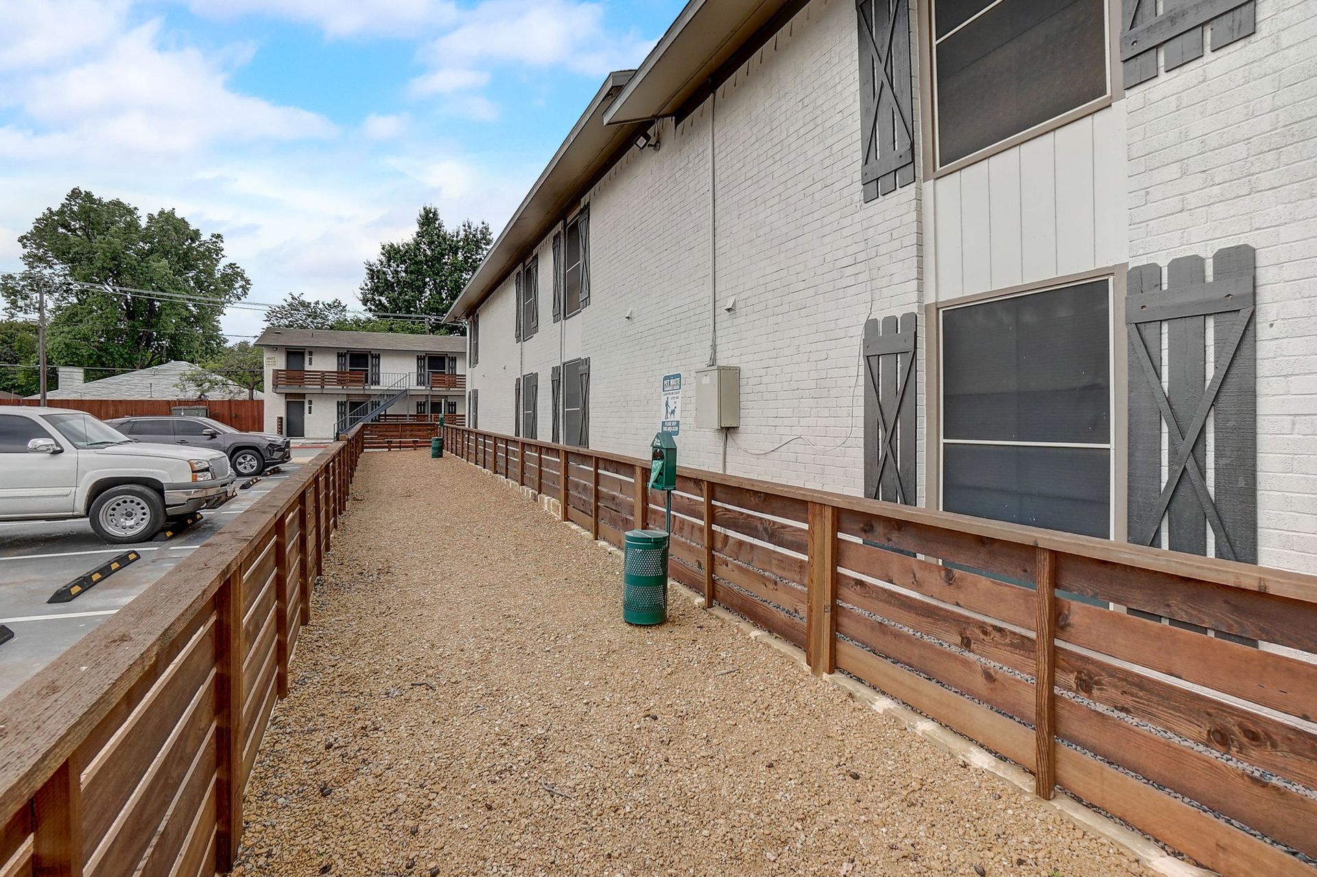 Dog park with gravel, wooden fence, and white brick apartment building.