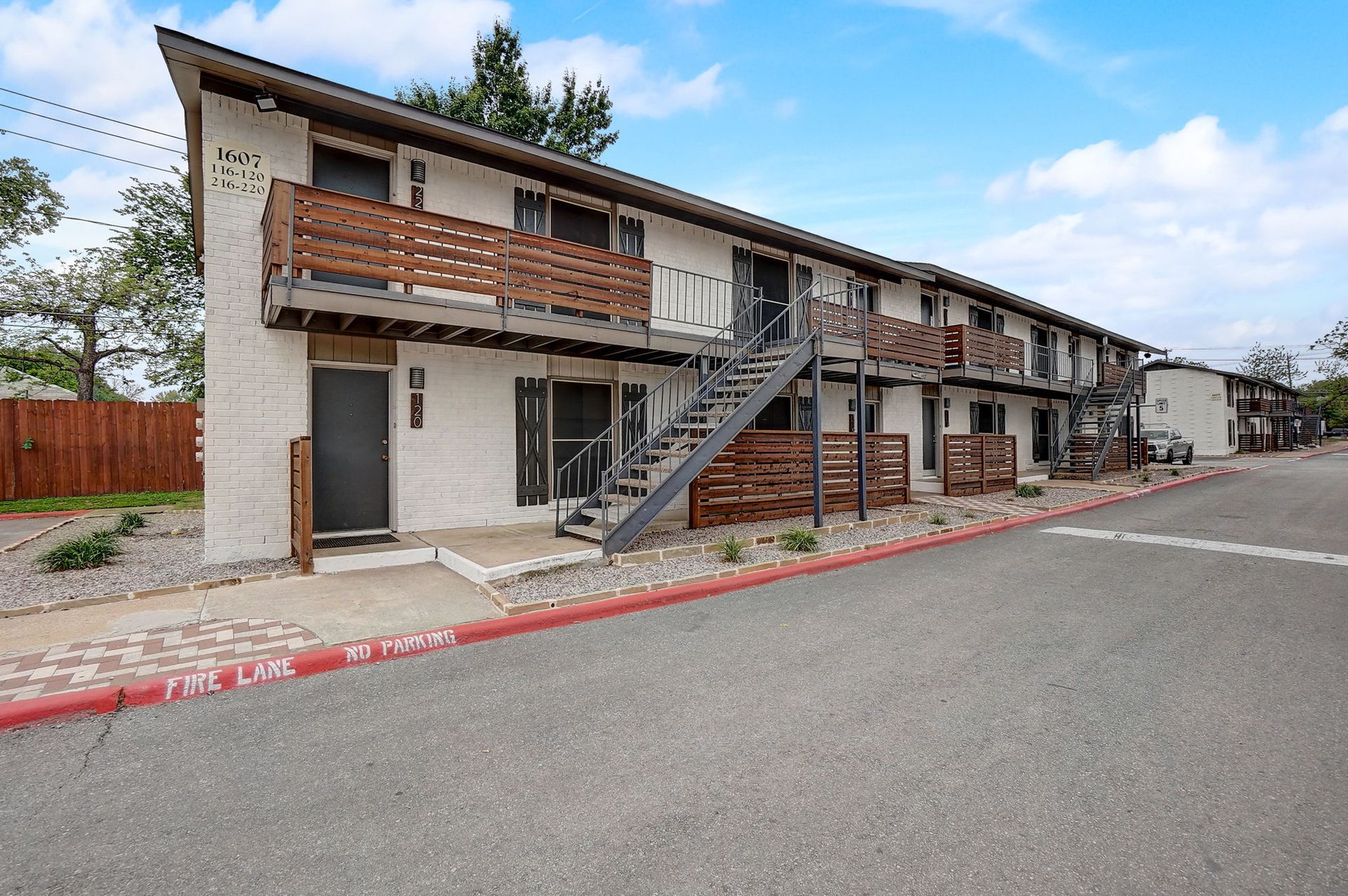 Apartment building with balconies and exterior staircase along a street under a blue sky.