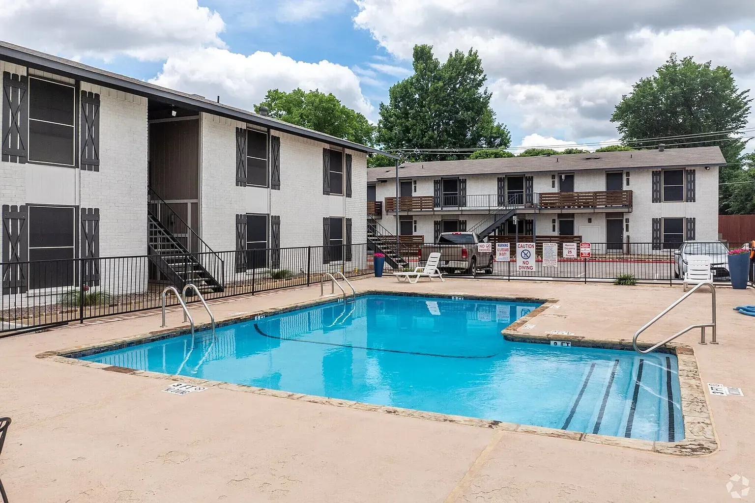 Apartment complex with a blue swimming pool in the foreground. Buildings are white with dark shutters.
