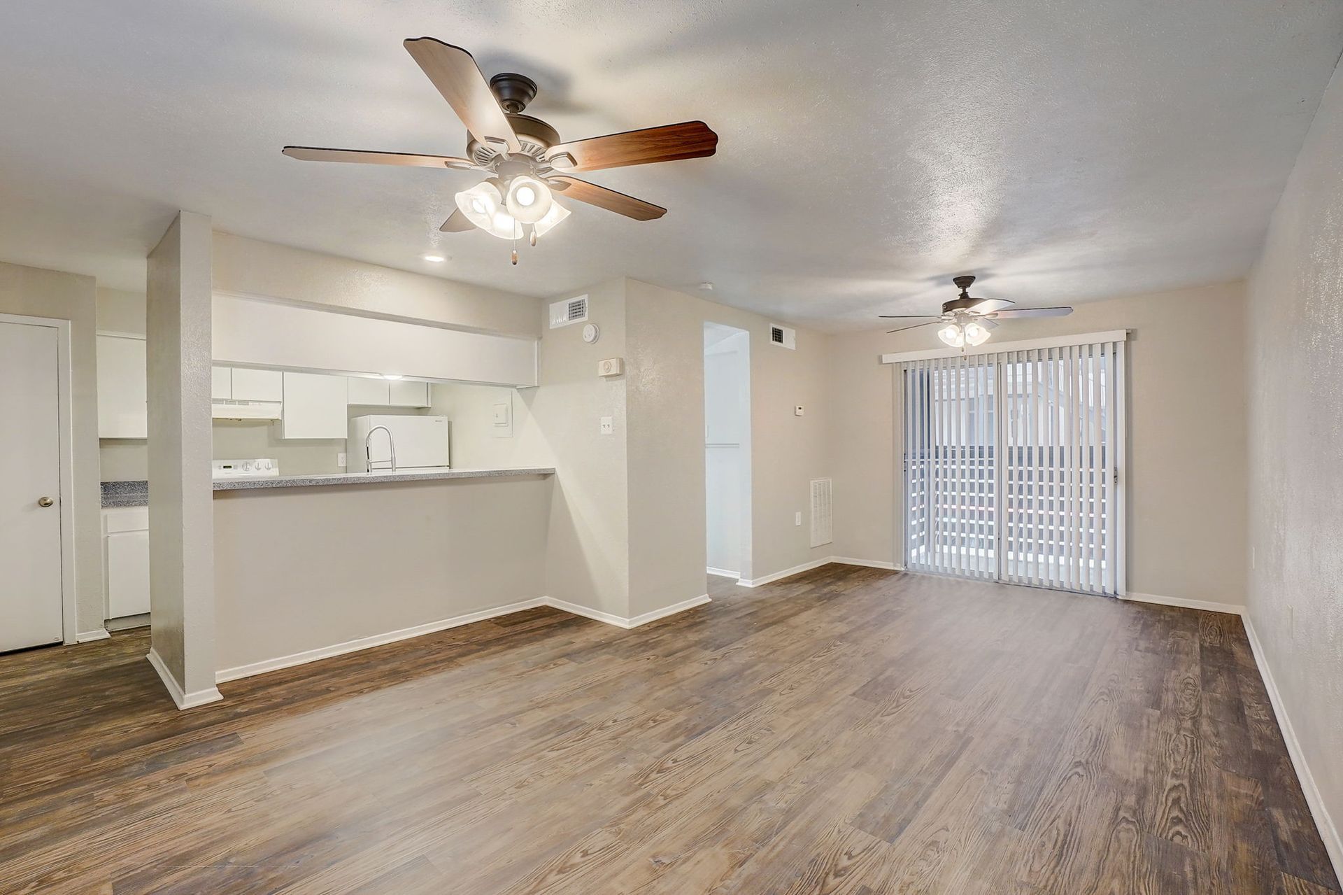 Empty living room with wood flooring, ceiling fans, and sliding glass door.