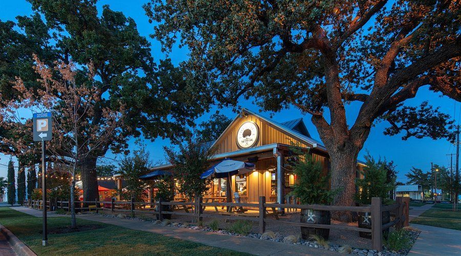 Rustic restaurant at dusk, under large trees with outdoor seating and a sign lit up.