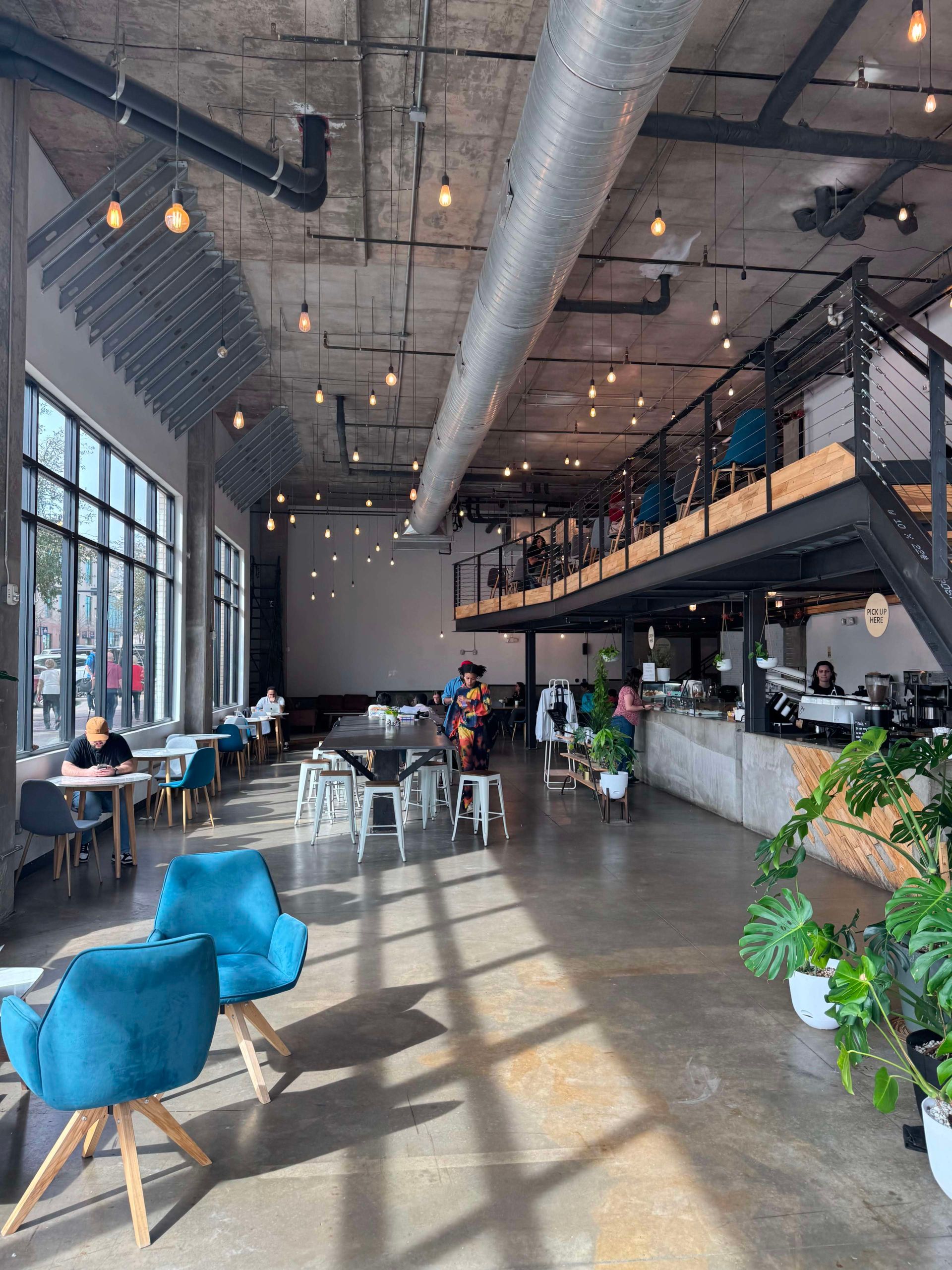 Interior of a modern cafe with high ceilings, featuring a mezzanine, seating, and large windows.