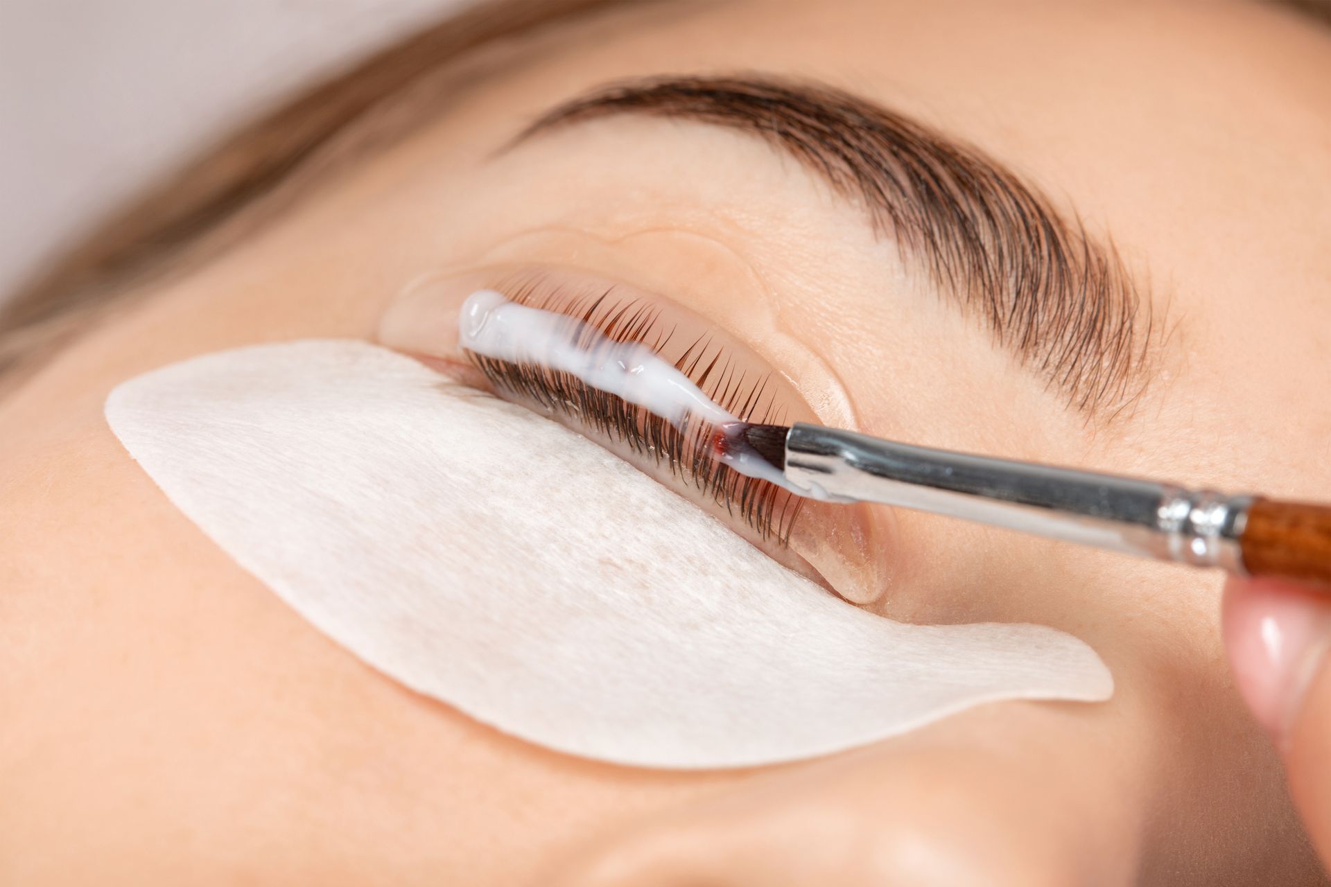 Person bei Wimpernlifting; Creme wird mit einem Pinsel auf die Wimpern aufgetragen. Augenklappe angelegt.