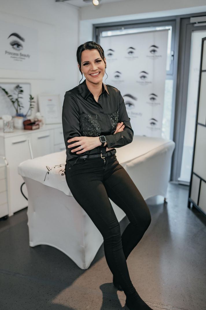 Eine Frau in schwarzem Hemd und Hose lächelt und lehnt sich in einem Schönheitssalon an eine Behandlungsliege.