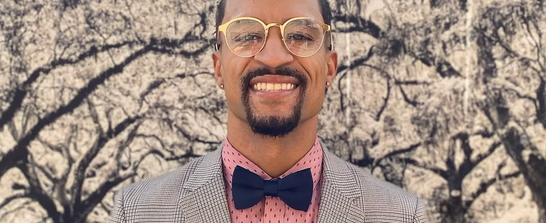 A man wearing glasses and a bow tie is smiling in front of a tree.