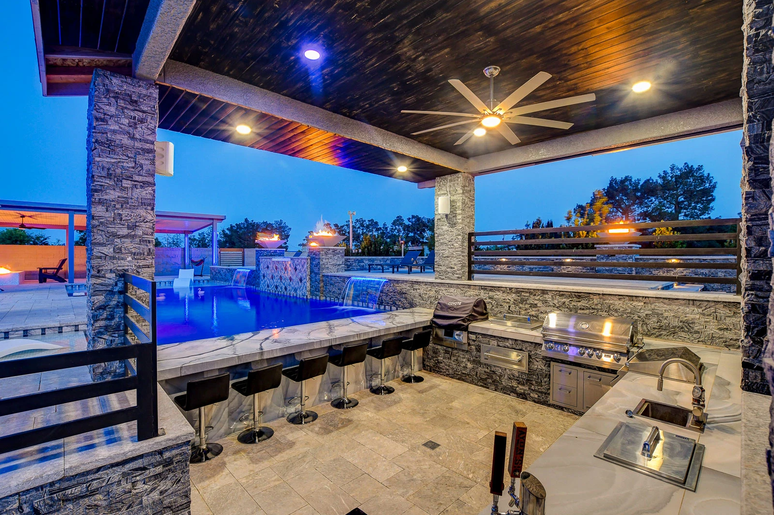 A large outdoor kitchen with a ceiling fan and a swimming pool in the background.