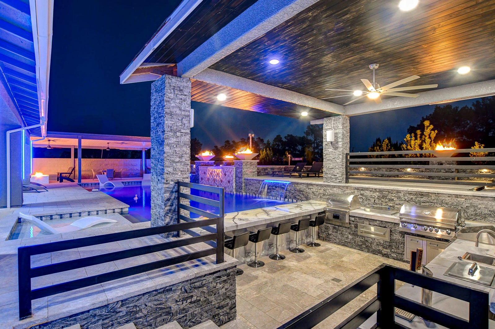 A large outdoor kitchen with a ceiling fan and a swimming pool at night.