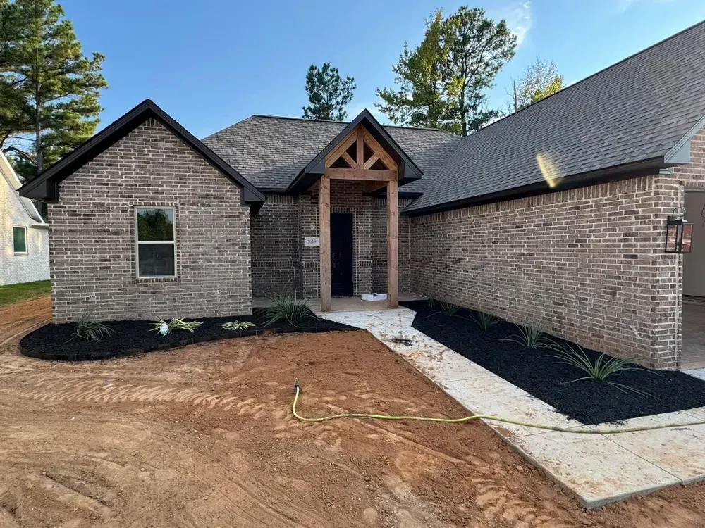 Brick house exterior with a dark roof and wooden porch detail, landscaping with black mulch.