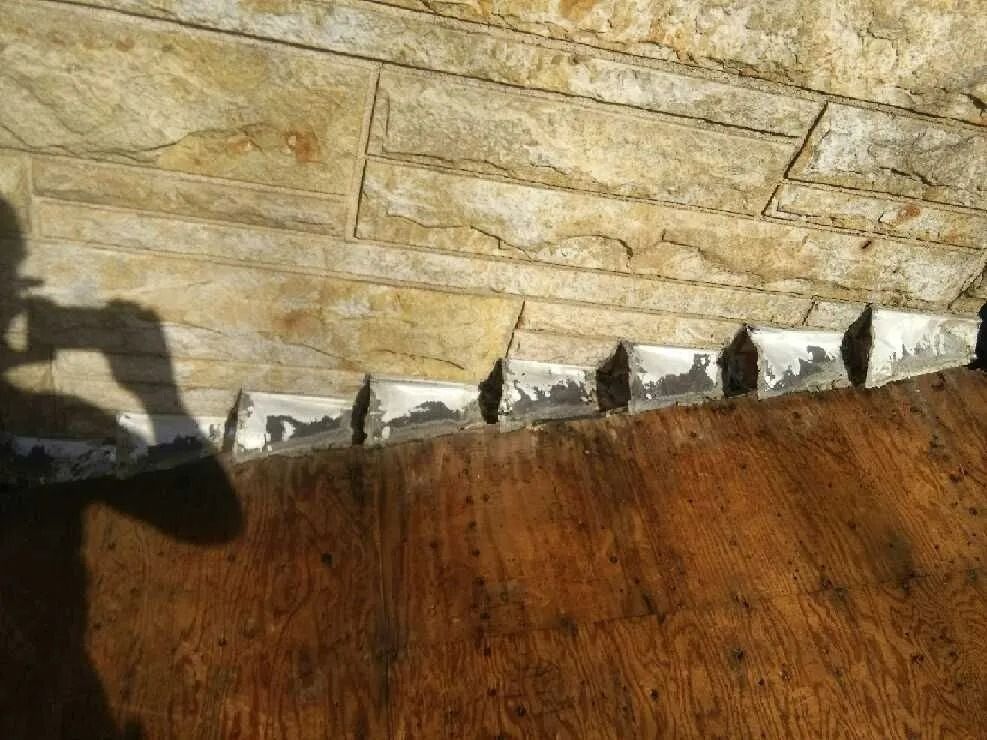 Stone veneer wall with peeling white sealant above a wood surface. Shadow of a person taking a photo.
