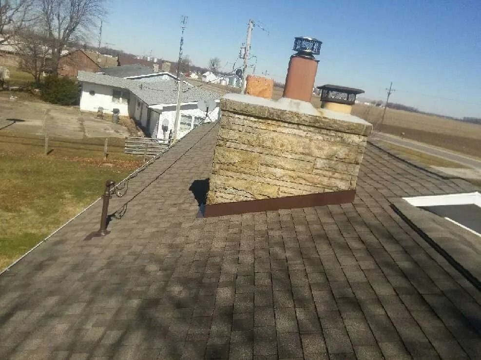 A brown shingle roof with a brick chimney and a small building in the background. Sunny day.