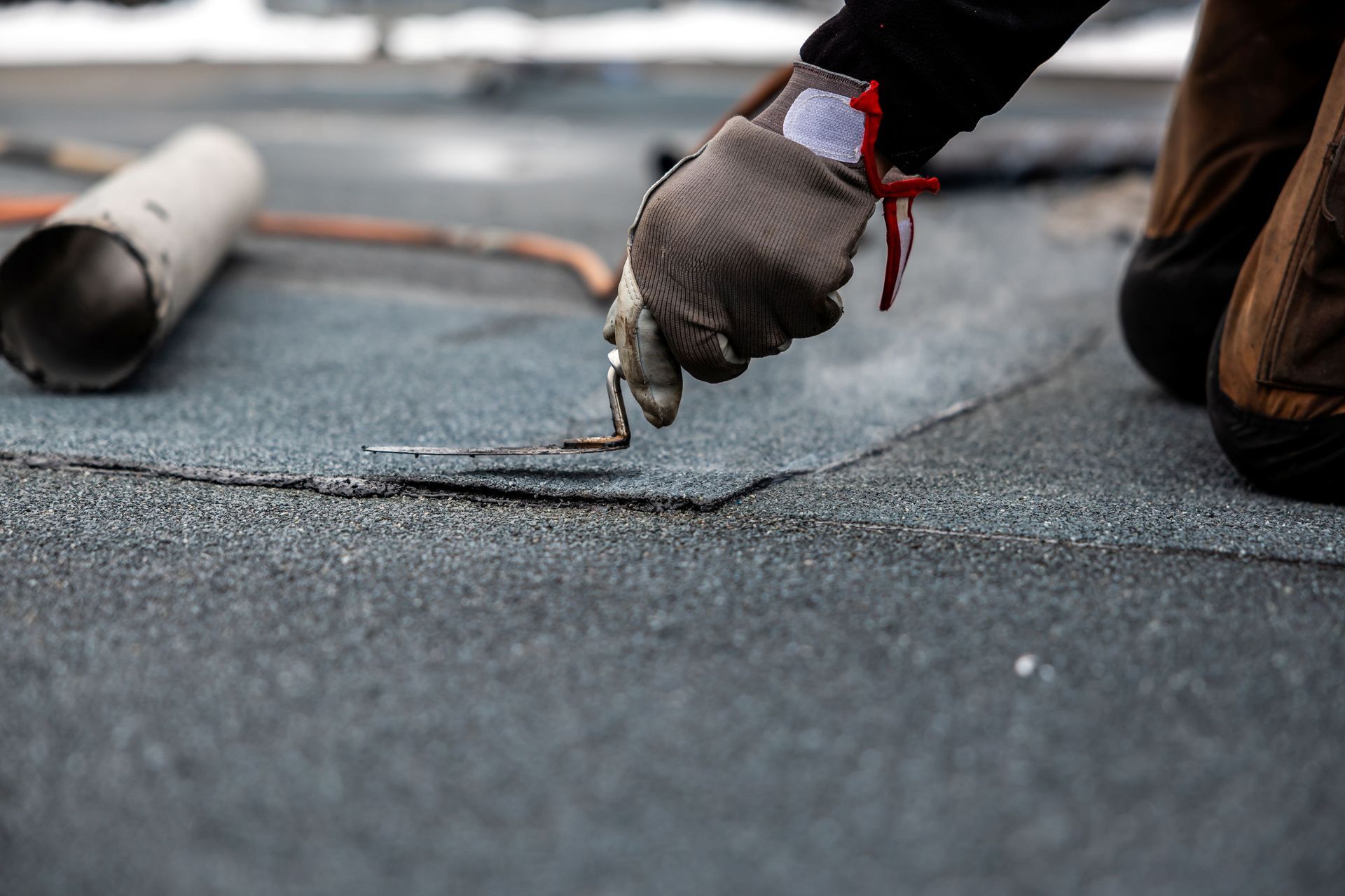 Person repairing a flat, dark roof with a hand tool, wearing gloves.