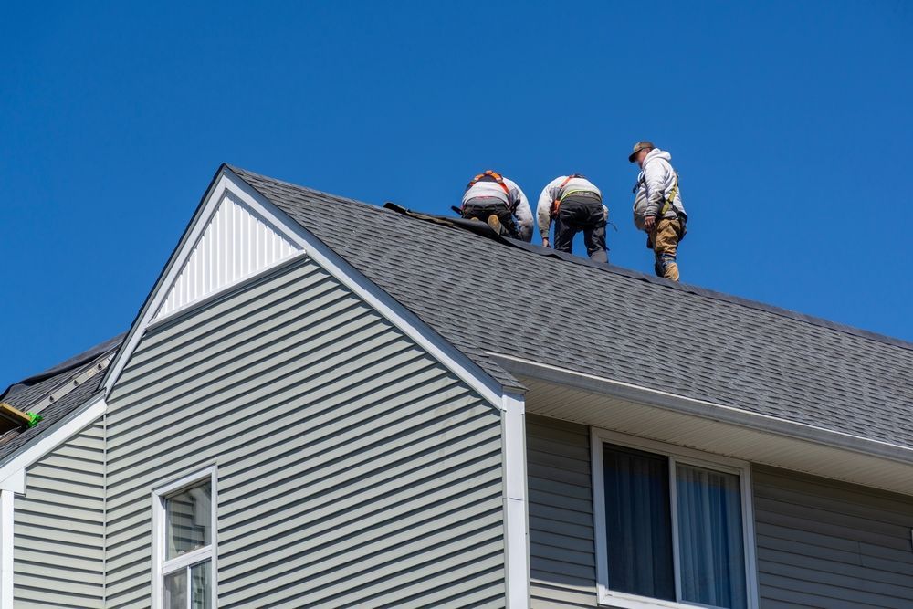 Three roofers working on a gray shingled roof, blue sky, white siding visible.