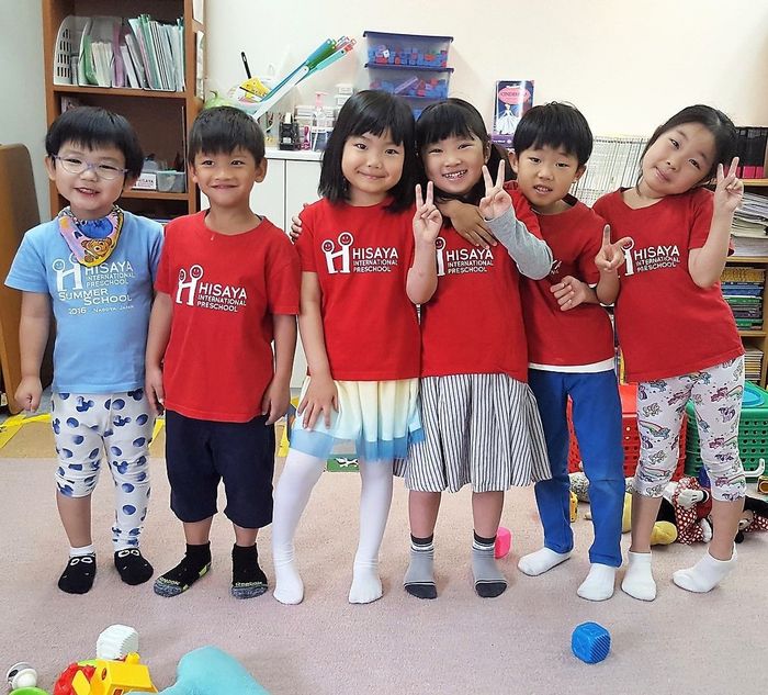 Six children posing indoors, smiling and holding up peace signs, wearing colorful shirts.