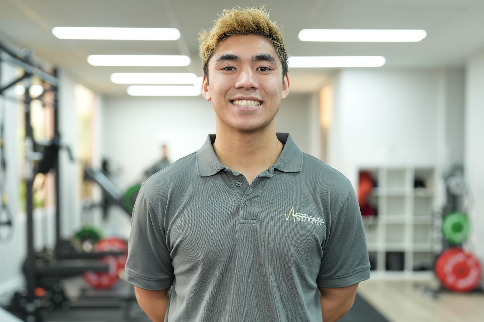 A young man in a grey polo shirt is standing in a gym and smiling.
