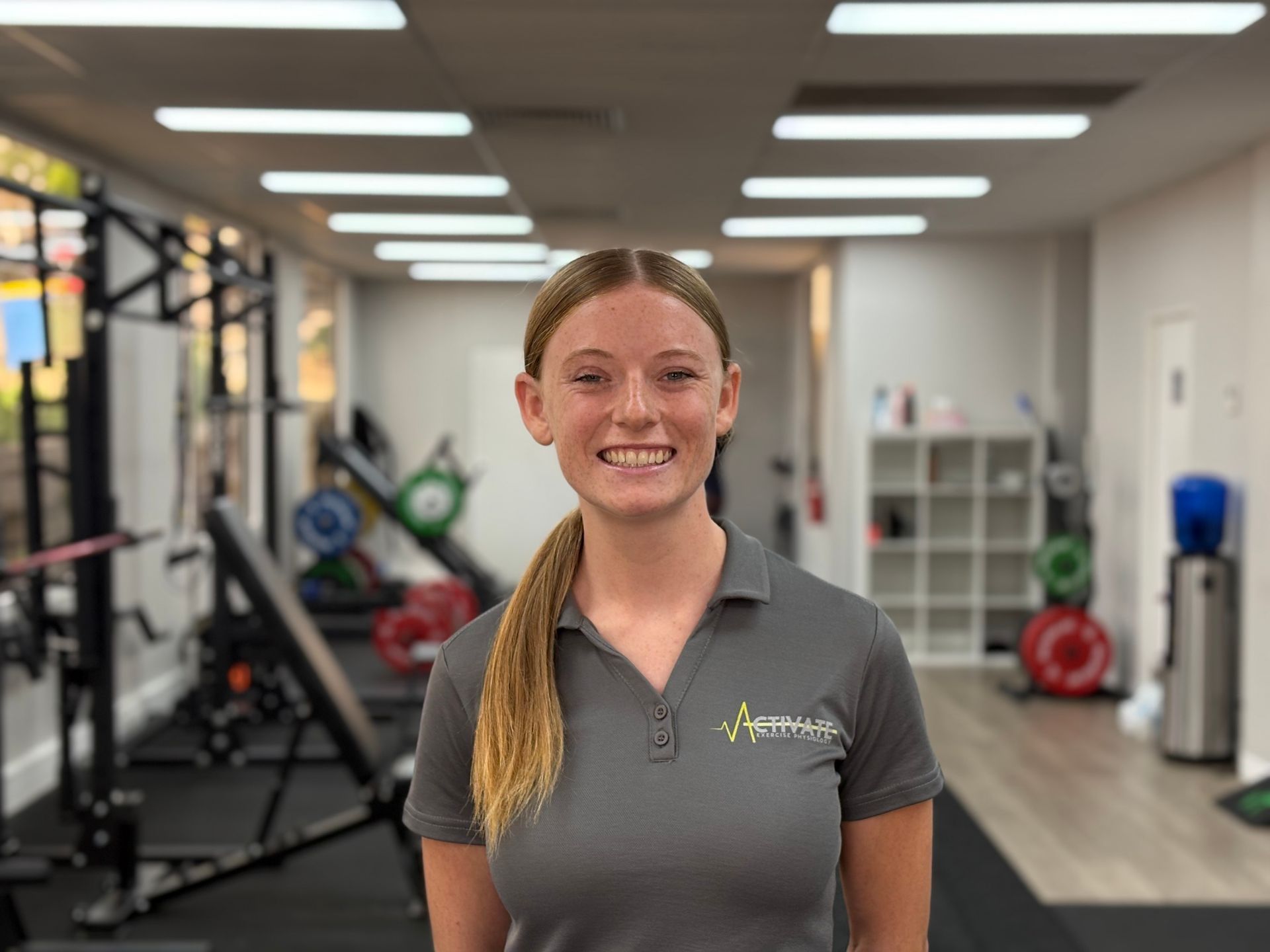 A woman is standing in a gym wearing a grey shirt and smiling.