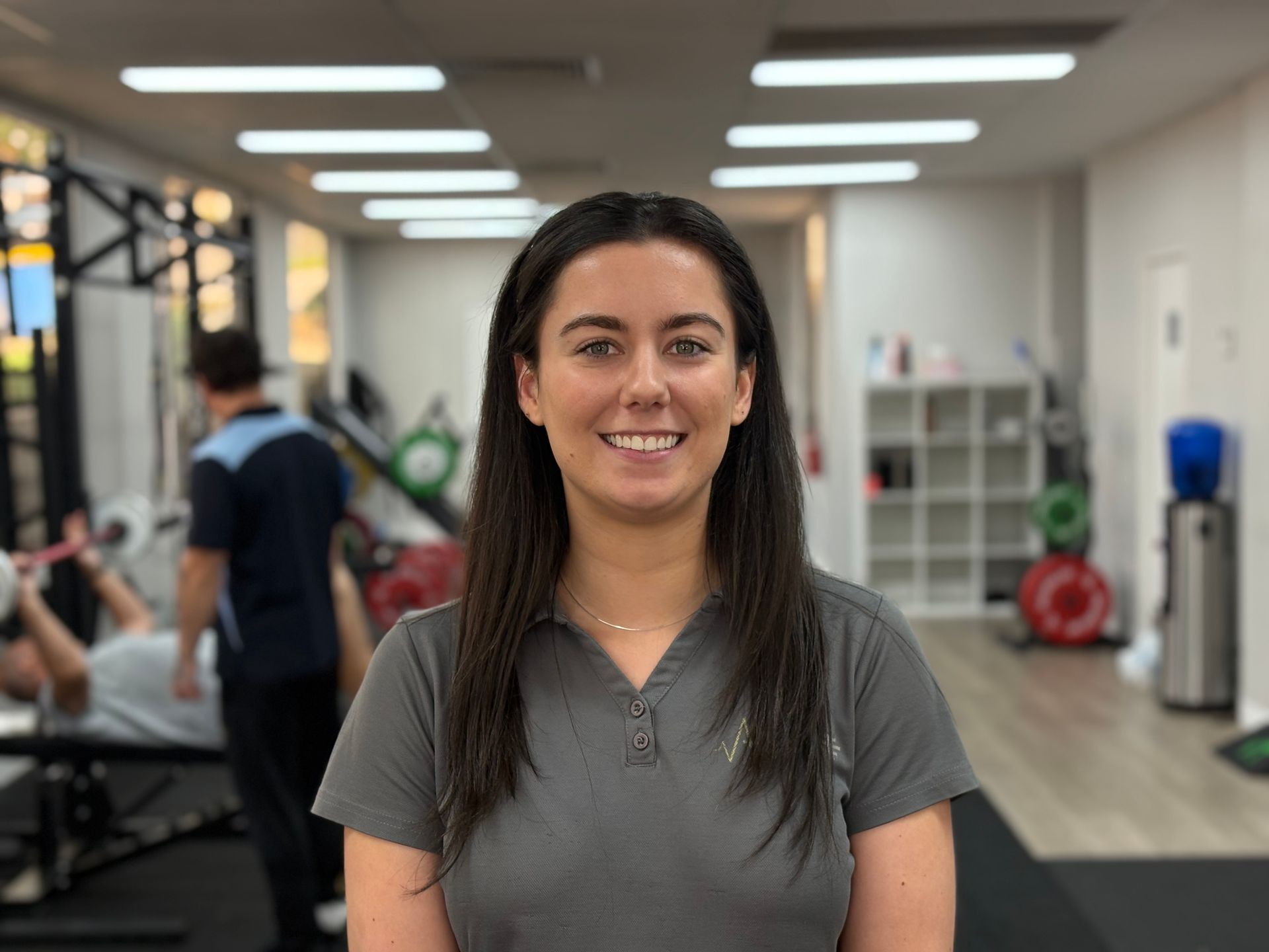 A woman is smiling in a gym with a man lifting weights in the background.