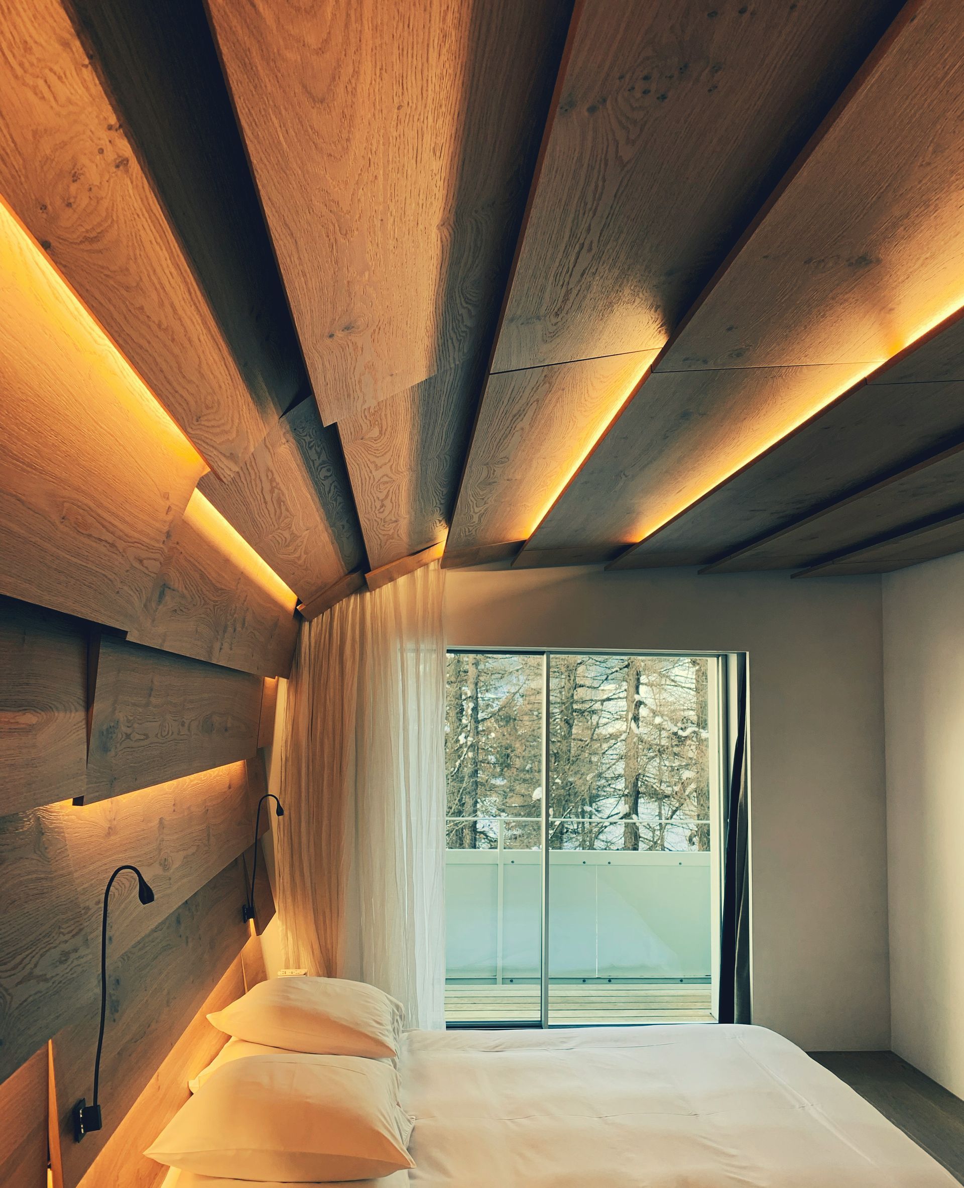 Bedroom with wooden ceiling and accent wall, warm lighting, and a balcony with trees.