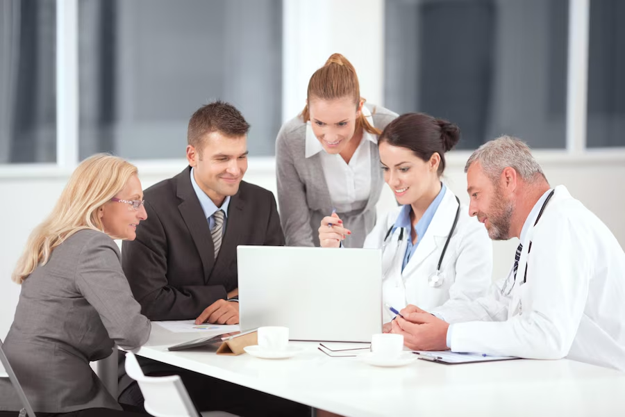 A group of doctors and business people are sitting around a table looking at a laptop.