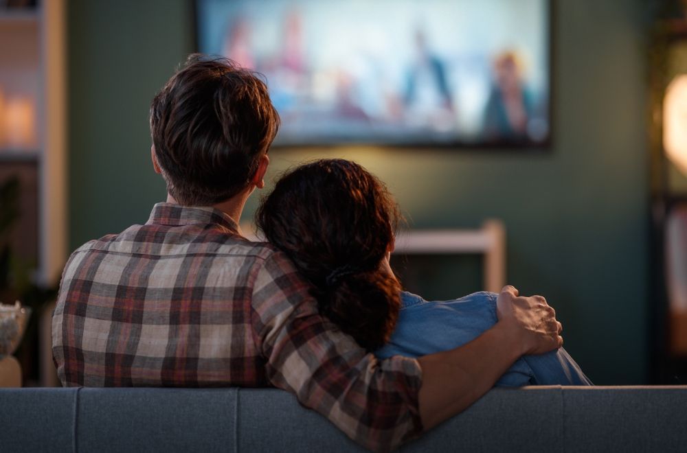 Couple on a sofa, watching a TV mounted on a green wall. Man's arm around woman, both looking at the screen.