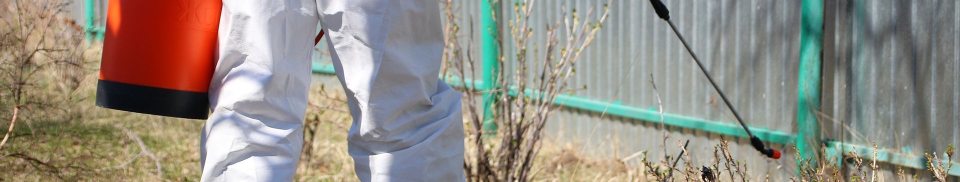 Person in white protective suit spraying weeds with an orange tank, next to a green fence.