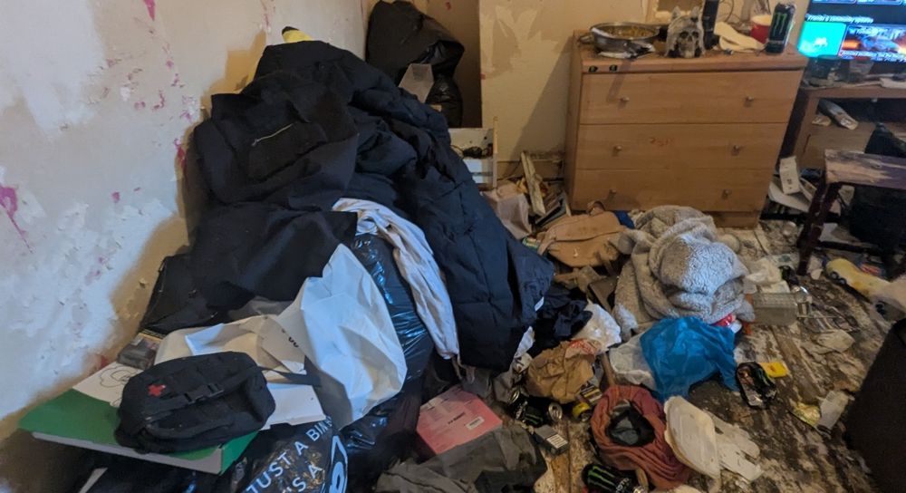 Messy room with clothes piled on the floor and against the wall, chest of drawers visible. before clean up