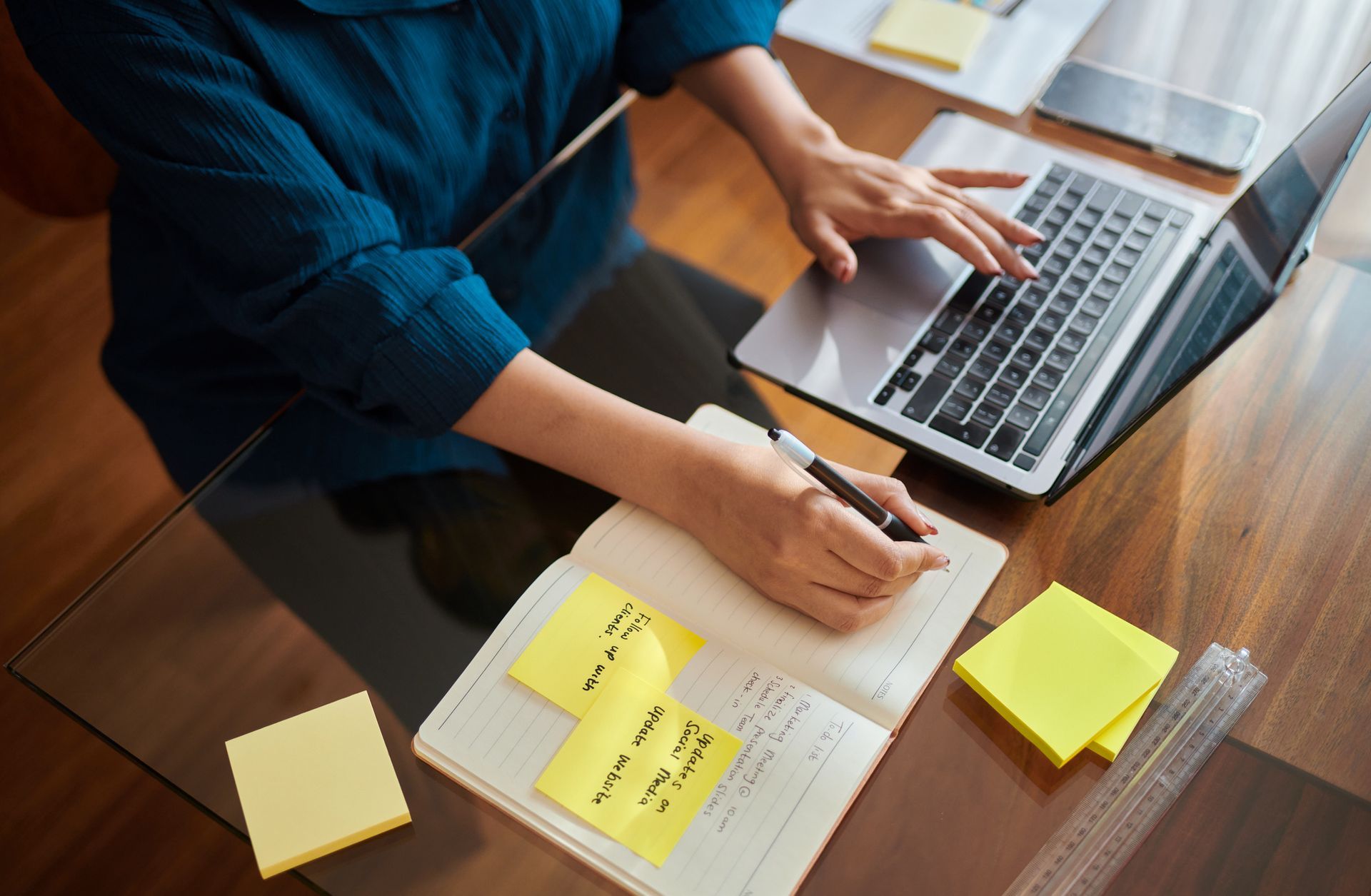 A woman is sitting at a desk using a laptop and writing in a notebook.