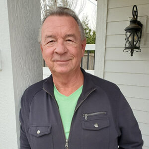 Man in gray jacket and green shirt smiles in front of a white building with a black lantern.