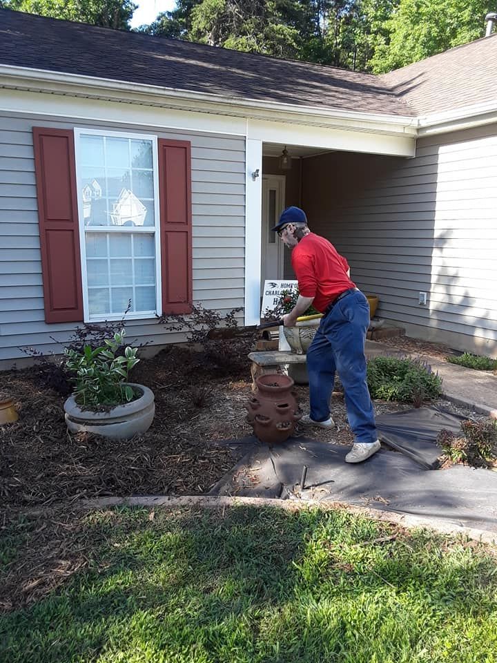 A person in a red shirt and blue jeans gardening in front of a house with light gray siding and red shutters.
