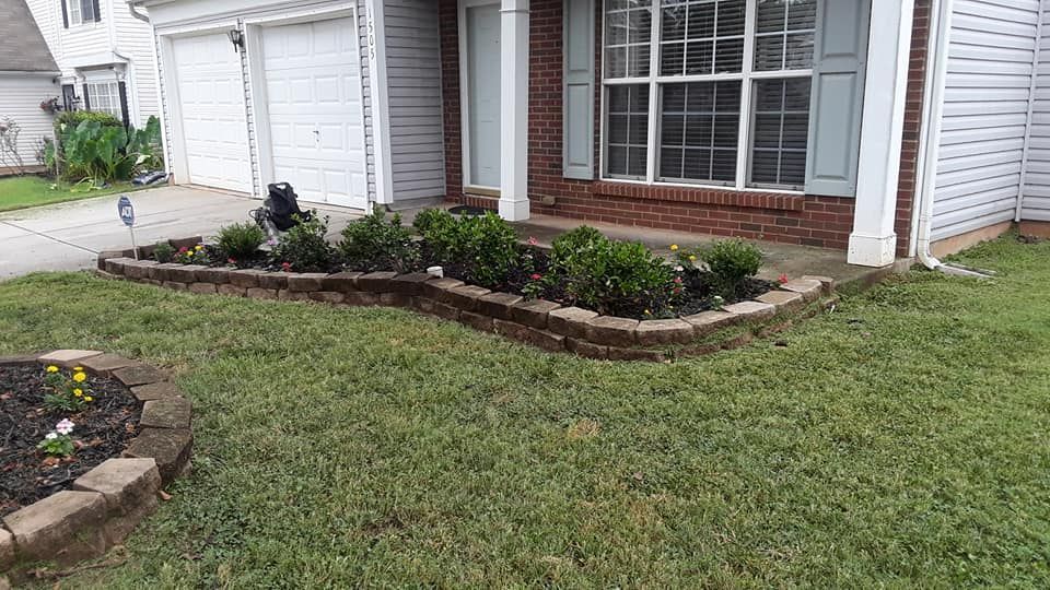 A raised flower bed with shrubs and a circular garden bed in front of a house with a brick facade and white siding.