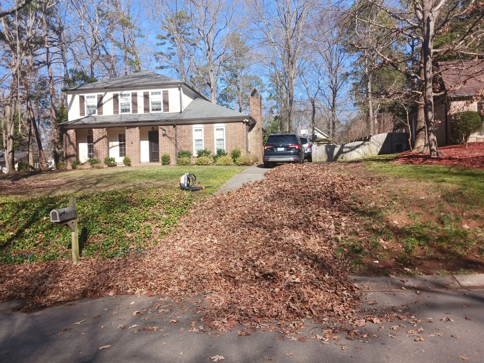 A two-story brick and white house sits on a suburban lot with a large pile of brown fallen leaves covering the driveway.