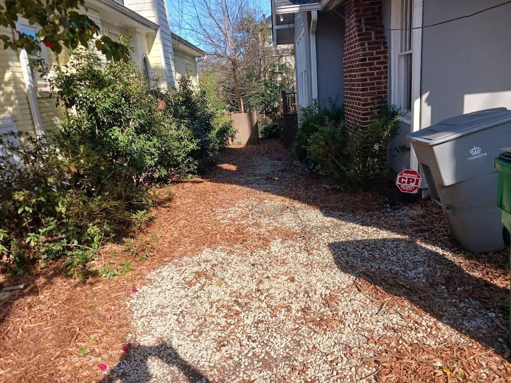 A side yard with mulch, a gravel pathway, dense greenery, a brick chimney, and a gray trash bin beside a light-colored house.