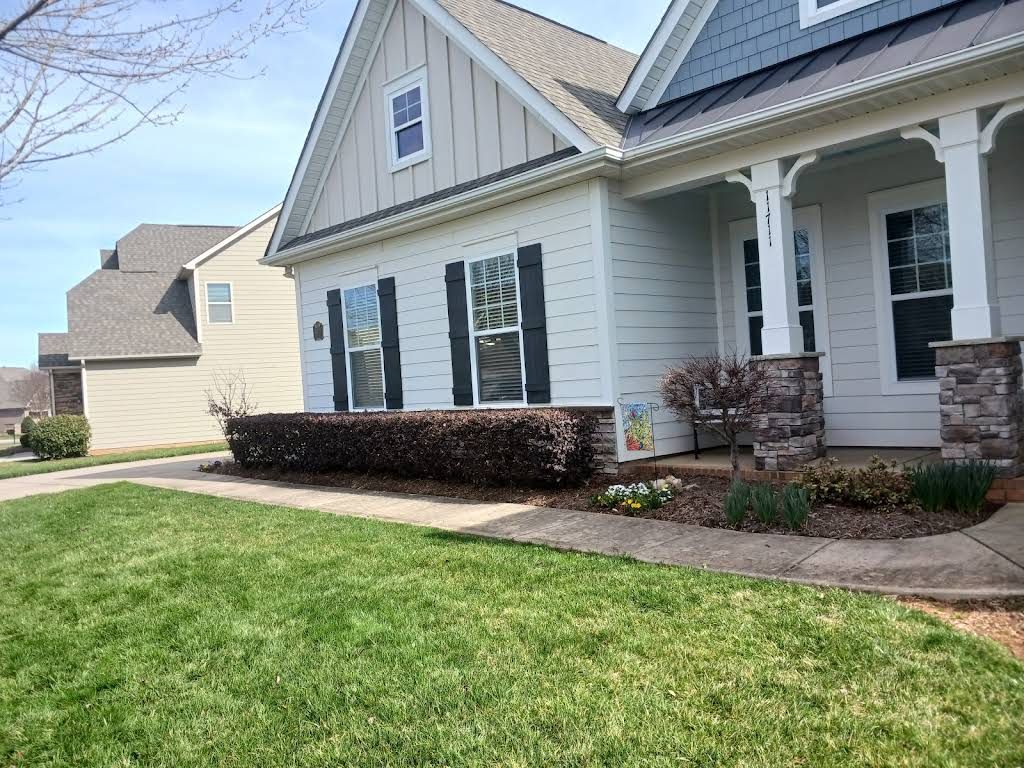 A white house with black shutters, a covered porch, and landscaped bushes in the front yard on a sunny day.